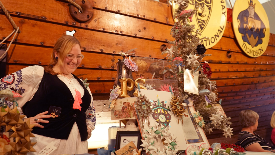 Image: a photograph showing local paper artist Nicki Marie in front of the Hjemkomst Viking ship. She smiles, wearing German clothing, standing next to her display booth of German ornaments and art.