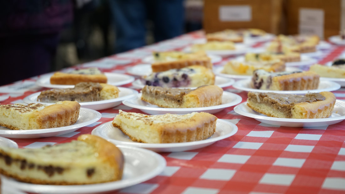 Image: a photograph showing several slices of Karen's Kuchen on a red/ and white checkered table. is seen in the foreground (drums, according, and keys) and the crowd sits and stands in the background, eating and drinking German fare.