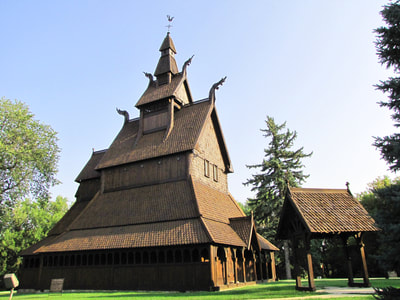 A long exterior shot of the Hopperstad Stave Church. The grass and trees around it are bright green, the sky in the background is cloudless and blue, and both the church and the gateway stand in the center of the photo. The 72-foot tall church looms large in the photograph.