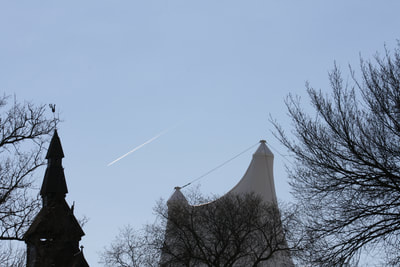 A long exterior shot of both the Hopperstad Stave church and the Hjemkomst Center. Bare tree branches stand in the foreground, the structures stand behind them, and a vapor jet trail cuts across the blue sky behind them.
