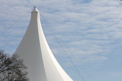 A long shot of the Hjemkomst Center, built in 1986 on the banks of the Red River in Viking Ship Park. The white Teflon tent stands tall in front of a blue sky and wisply clouds. 
