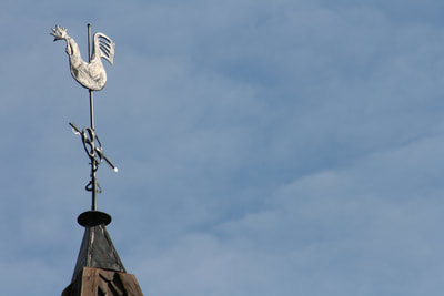 A closeup exterior shot of the Hopperstad Stave Church weather vane: a rooster on a steel pole at the top of the church. A blue sky is seen in the background.