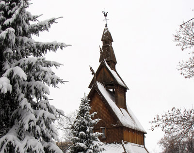 A long exterior shot of the Hopperstad Stave Church taken in the winter. A Douglas Fir stands in the foreground, framing the left of the picture, another smaller fir stands in the center, and the church looms in the background. The roofs are covered with snow and the weather vane is seen at the top.