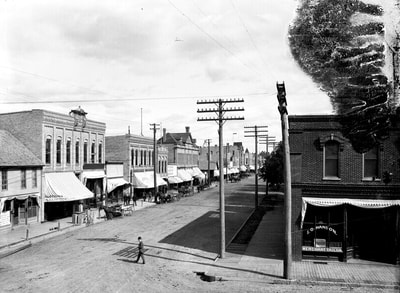 A black & white exterior shot of Moorhead taken by O.E. Flaten circa 1895. The image faces eastward from the corner of Center Avenue and 4th Street, showing the old downtown brick buildings and a man in a bowler hat crossing the street.