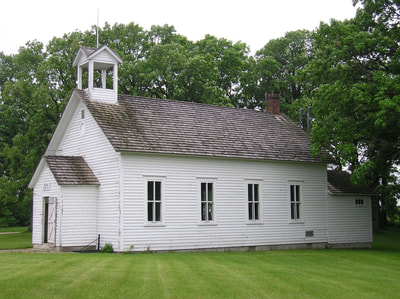 A long exterior shot of the Woodlawn School. This historic building used to serve rural students until consolidation brought them to Moorhead. The building is rectangular with a door on the end, four windows on the side, white siding, gray shingles, and a bell cupola at the top. 