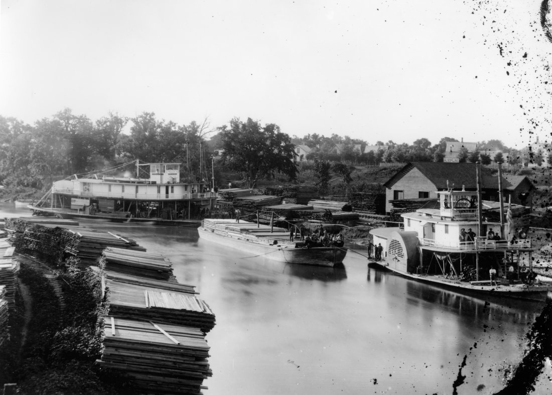 Image: an 1881 photograph of the Fargo-Moorhead riverfront. Steamboats and barges line the banks.