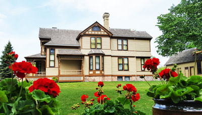 A long exterior shot of the Comstock House in south Moorhead. Bright red roses and the green lawn stretch out in the foreground and the Queen Anne Victorian mansion stands in the background with a yellow-beige siding, large windows, and wood tone trim.