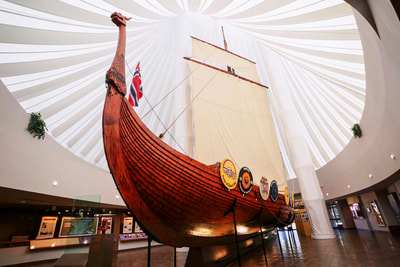 A wide-angle photograph of the Hjemkomst, our namesake Viking ship. The dragon masthead Igor stands tall on the left, the ship stretches back and to the right, and the mast stands tall in the center.