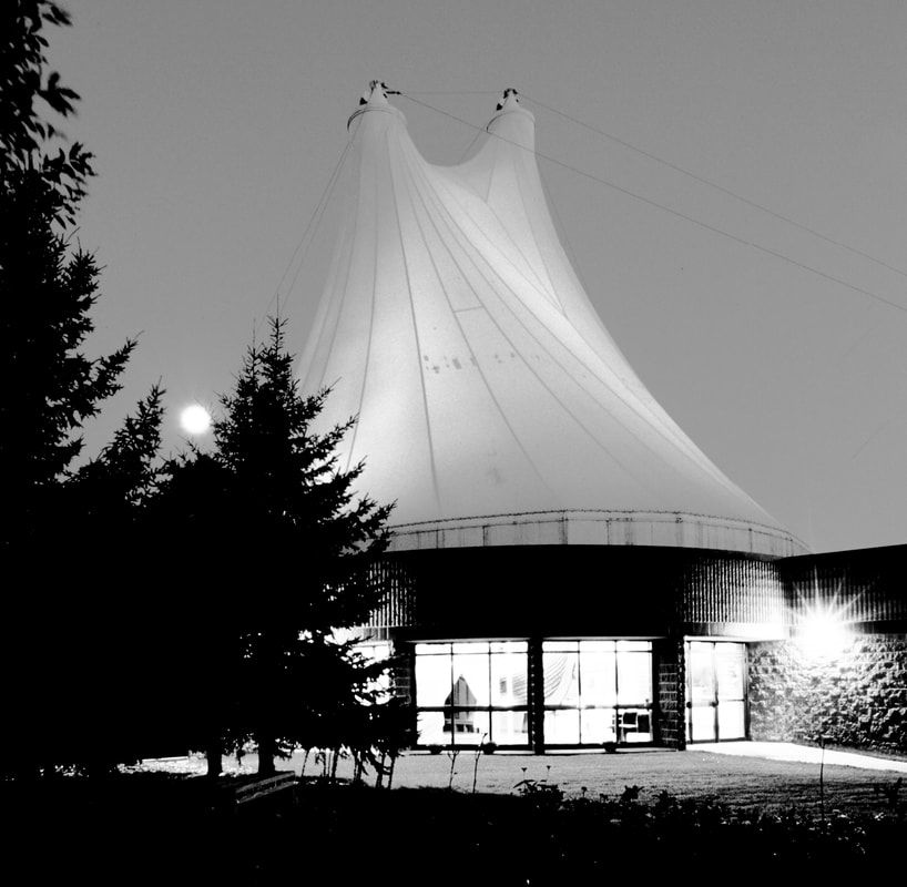Image: a black and white exterior photograph of the Hjemkomst Center. The white teflon roof stretches to the sky. The moon and interior lights shine through pine trees outside.